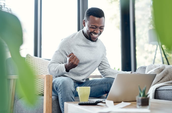 A man sits, looking at a laptop, smiling and clenching his fists.